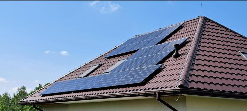 Panele fotowoltaiczne zamontowane na brązowym dachu skośnym z dachówką ceramiczną, widoczne okna dachowe i antena.