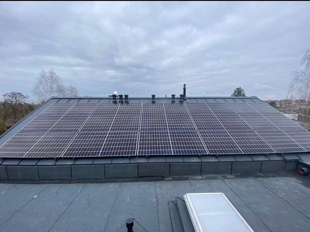 Panele słoneczne na dachu budynku z szarej blachy w pochmurny dzień, widoczne kominy i elementy wentylacyjne.