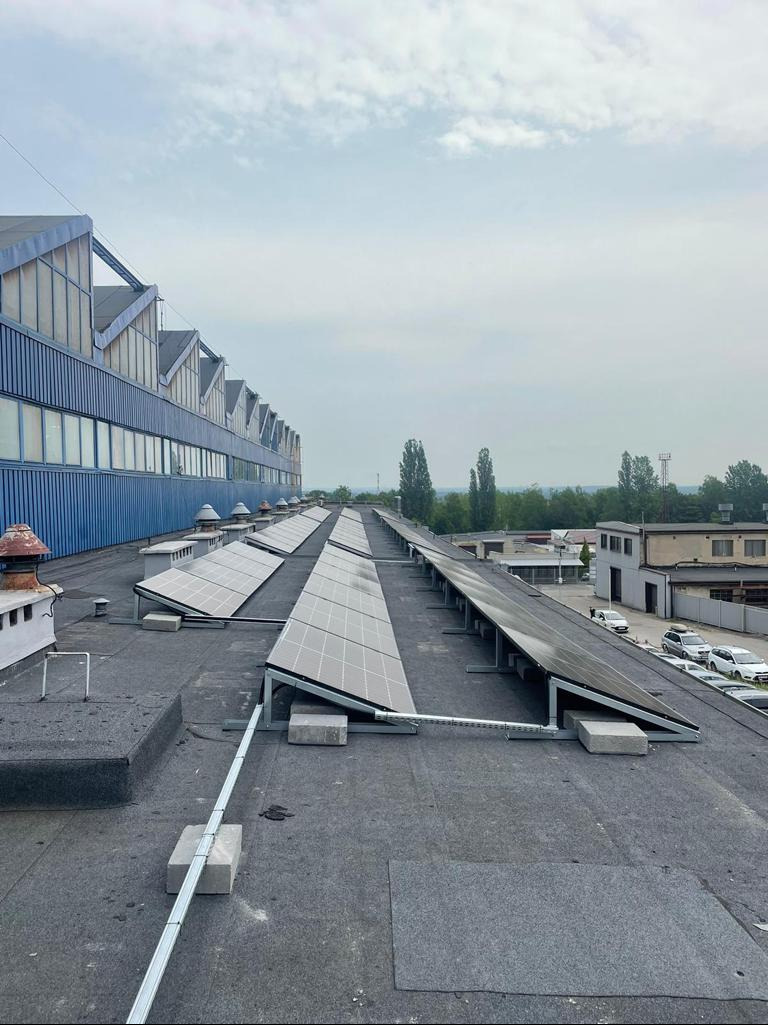 Rząd paneli słonecznych na dachu budynku przemysłowego, widoczne betonowe bloki podtrzymujące konstrukcję, w tle zabudowania i drzewa.