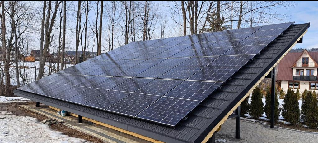 Panel fotowoltaiczny na dachu z blachodachówki, widok na dom w tle, częściowo ośnieżony teren.