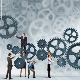 Zespół w kaskach i garniturach montuje system zębatych kół na tle abstrakcyjnego wzoru zębatek.