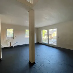 Wnętrze w trakcie prac wykończeniowych w Kamieniu Pomorskim: świeżo wylana, czarna wylewka podłogowa i otynkowane ściany w surowym stanie, z widocznymi instalacjami.