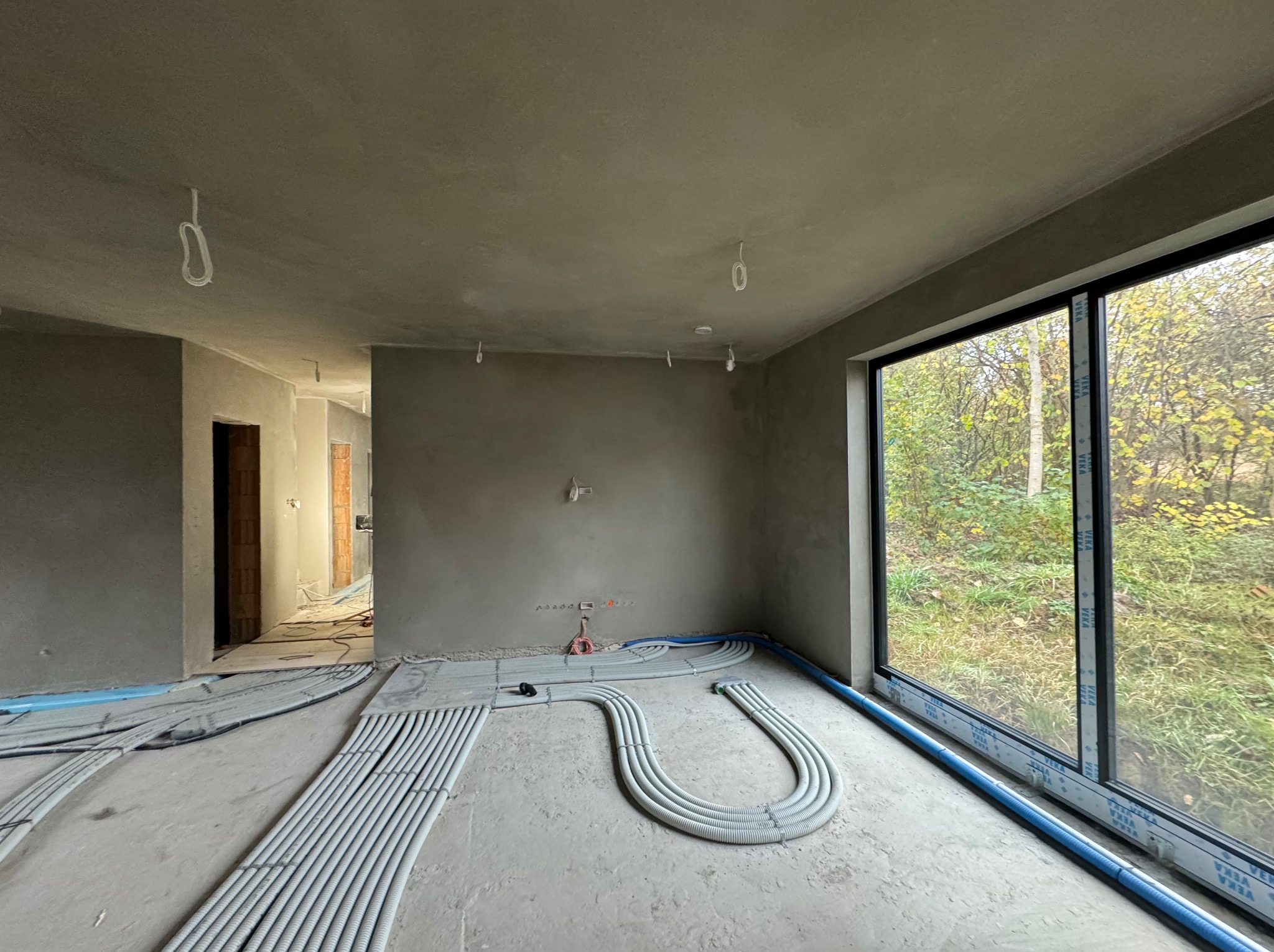 Wnętrze w stanie deweloperskim po tynkach maszynowych, z widocznymi przewodami elektrycznymi w peszlach na podłodze i dużym oknem z widokiem na jesienny ogród.