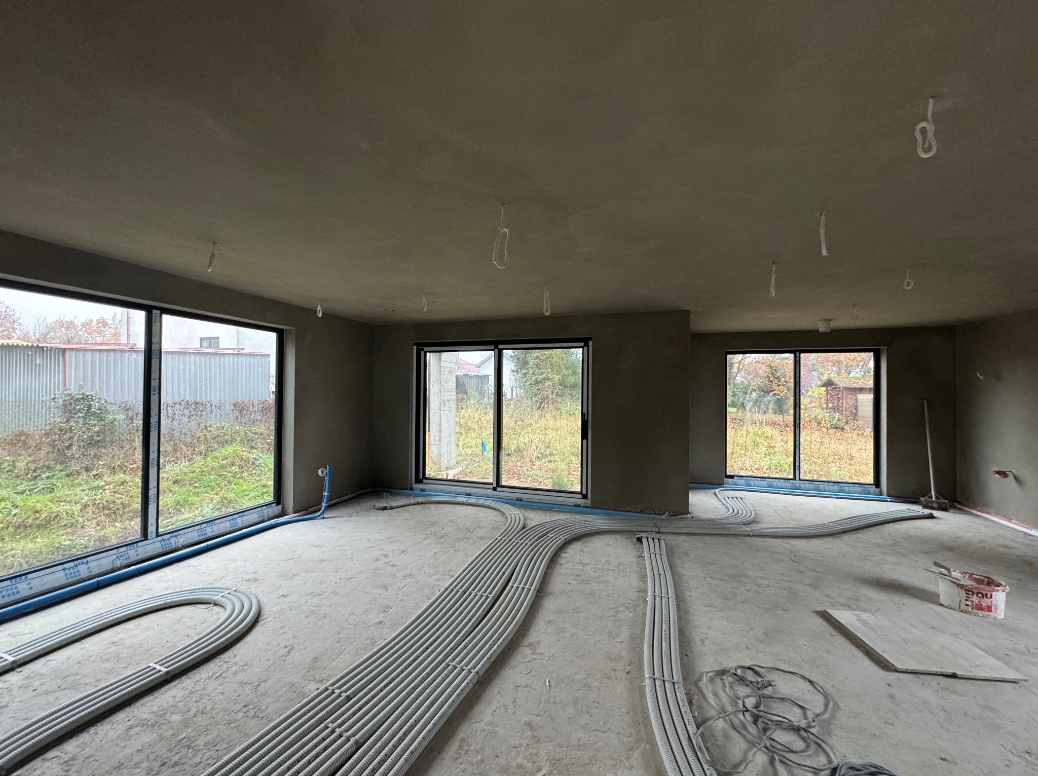 Surowe wnętrze po nałożeniu szarego tynku maszynowego, z widocznymi instalacjami elektrycznymi w peszlach na podłodze i wystającymi przewodami z sufitu, duże okna z widokiem na zewnątrz.