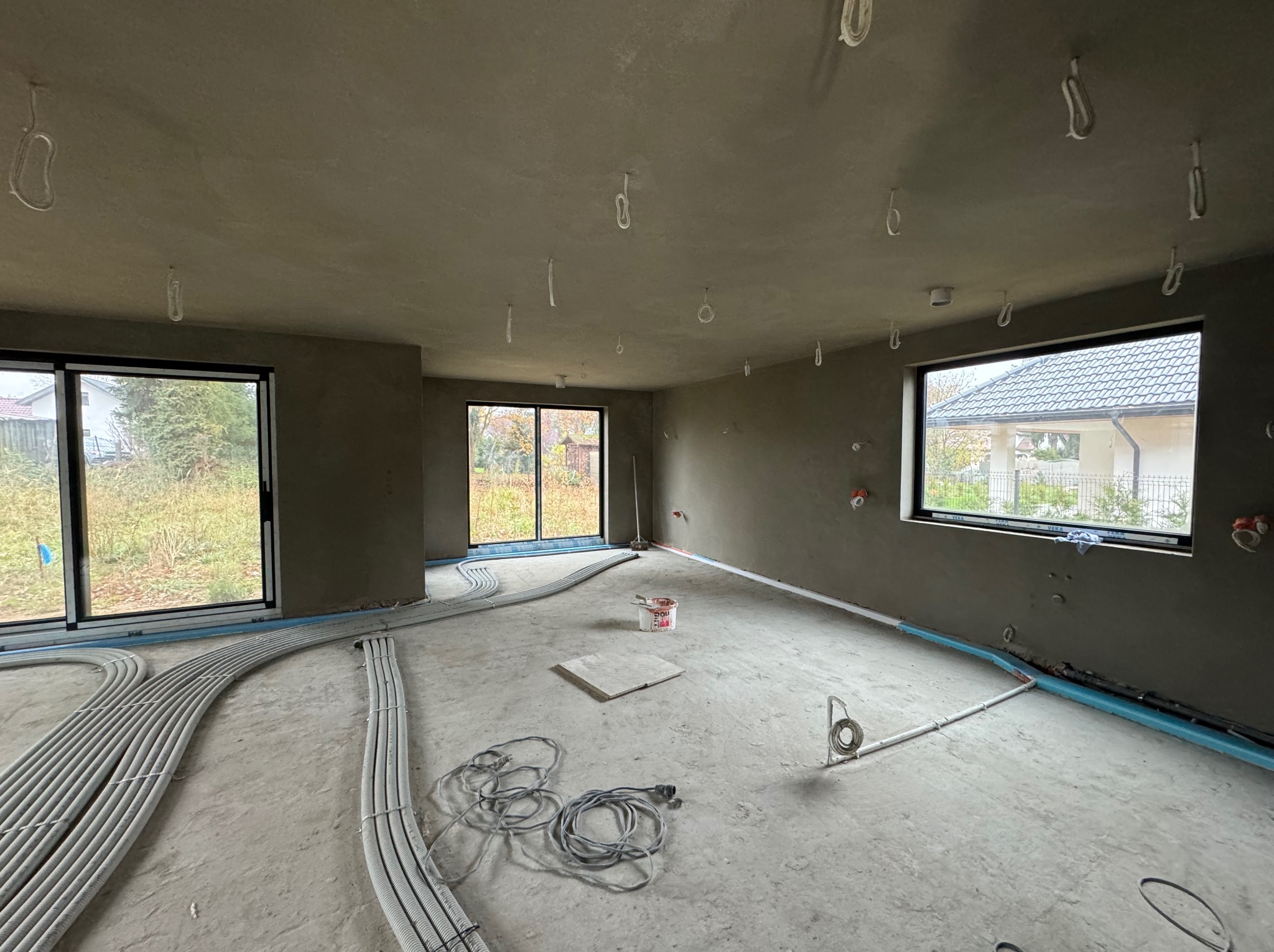 Wnętrze surowego domu po nałożeniu tynków maszynowych, widoczne kable elektryczne zwisające z sufitu oraz rury ogrzewania podłogowego wzdłuż ścian, przygotowane do dalszych prac wykończeniowych.