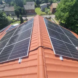 Widok z góry na panele słoneczne na czerwonym dachu domu jednorodzinnego, z widocznymi elementami montażowymi i otoczeniem zieleni.