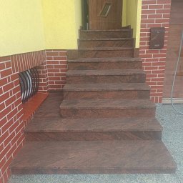 Kamieniarstwo BORSTONE (KAM-BOR) - Solidne schody wejściowe z brązowego granitu, prowadzące do drewnianych drzwi. Obok żółta ściana z tynkiem barankowym i elewacja z czerwonej cegły klinkierowej z brązową skrzynką pocztową.