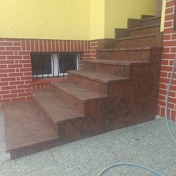 Kamieniarstwo BORSTONE (KAM-BOR) - Zewnętrzne schody z polerowanego, brązowo-czerwonego granitu, z widocznym wzorem, przy żółtej elewacji i ceglanej ścianie z oknem z kratą.