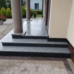 Kamieniarstwo BORSTONE (KAM-BOR) - Mokre, dwukolorowe schody wejściowe z jasnoszarego i ciemnego granitu, prowadzące do pasującego podestu pod dwoma filarami. W tle zielone krzewy i dom.
