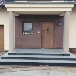 Kamieniarstwo BORSTONE (KAM-BOR) - Wejście do domu z trzema stopniami z jasnoszarego kamienia i ciemnymi podstopnicami, podtrzymywane przez fakturowane beżowe kolumny. Drewniane drzwi wejściowe i ceglana elewacja.