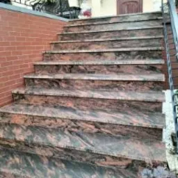 Zewnętrzne schody z ciemnego kamienia z wyraźnym, nieregularnym wzorem, prowadzące do wejścia do budynku. Widoczna ceglana ściana po lewej stronie i metalowa balustrada po prawej.