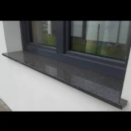 Zamontowany, ciemny, granitowy parapet zewnętrzny pod oknem z szarą ramą, widoczny fragment elewacji budynku.