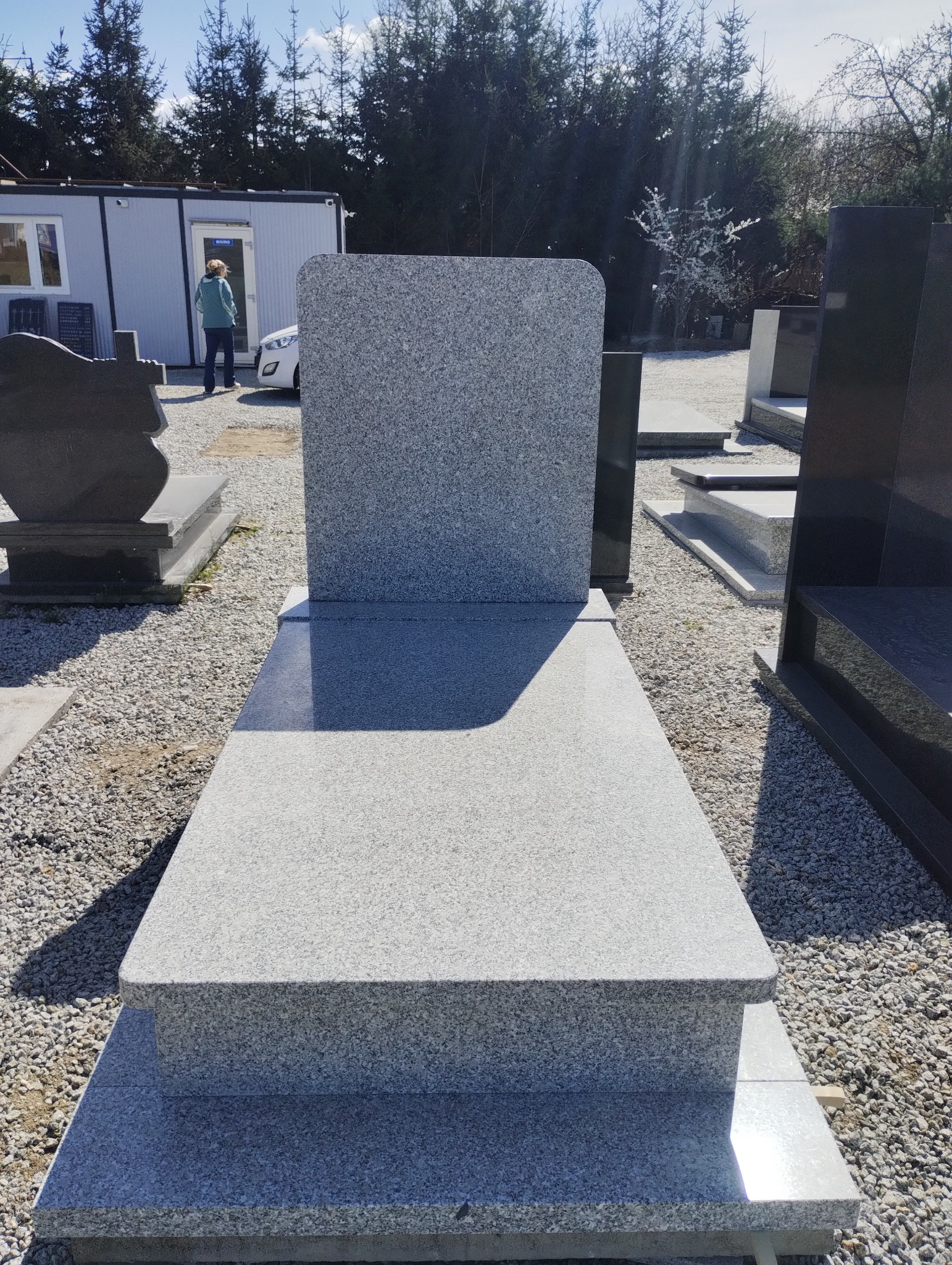 Jasnoszary, granitowy pomnik nagrobny z zaokrąglonymi górnymi rogami, ustawiony na żwirowym terenie, z widocznym budynkiem biurowym i drzewami w tle.