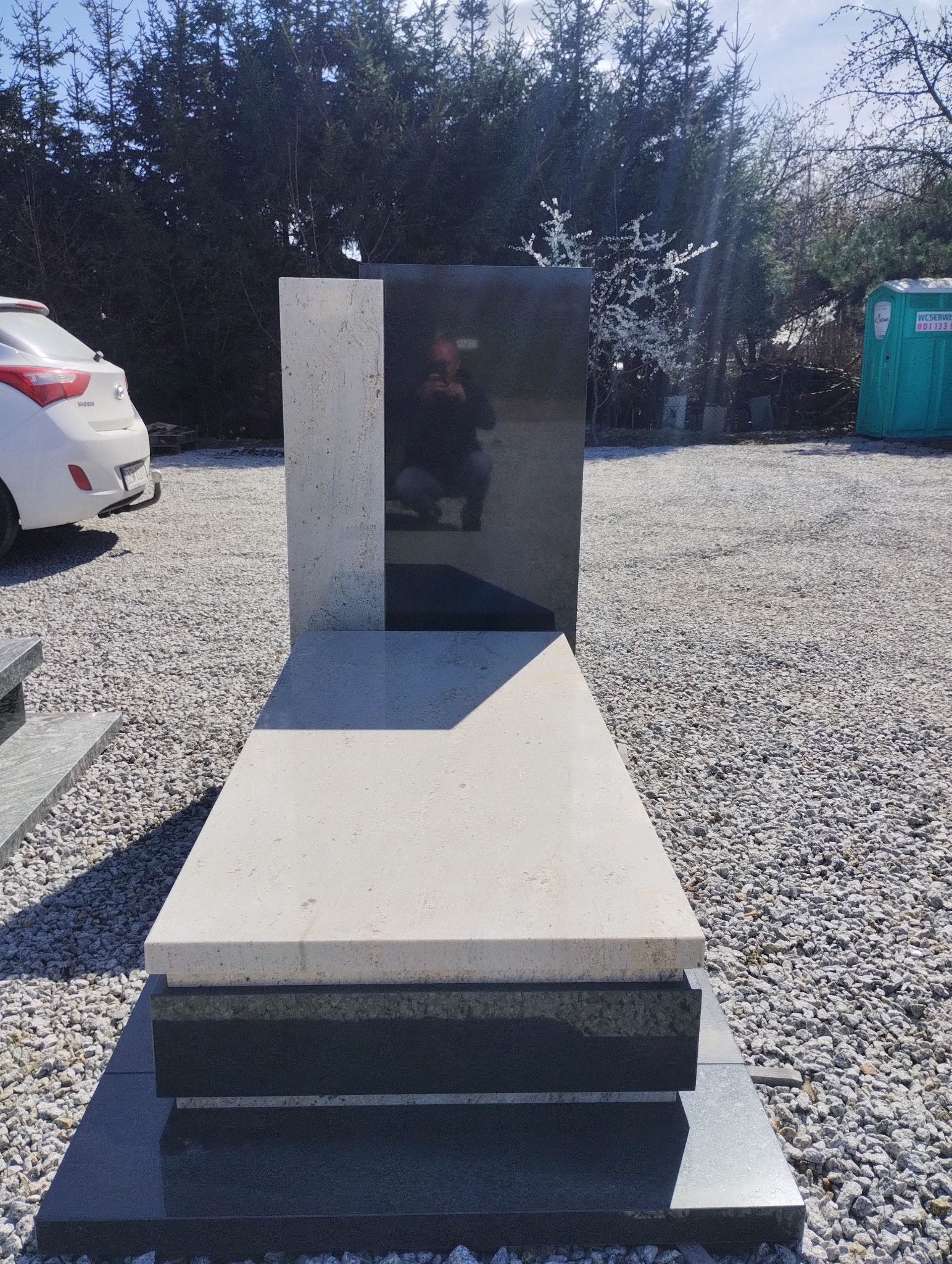 Nowoczesny nagrobek z jasnego i ciemnego kamienia, z odbiciem fotografa w polerowanej powierzchni, ustawiony na żwirowym terenie z widocznym samochodem i przenośną toaletą w tle.