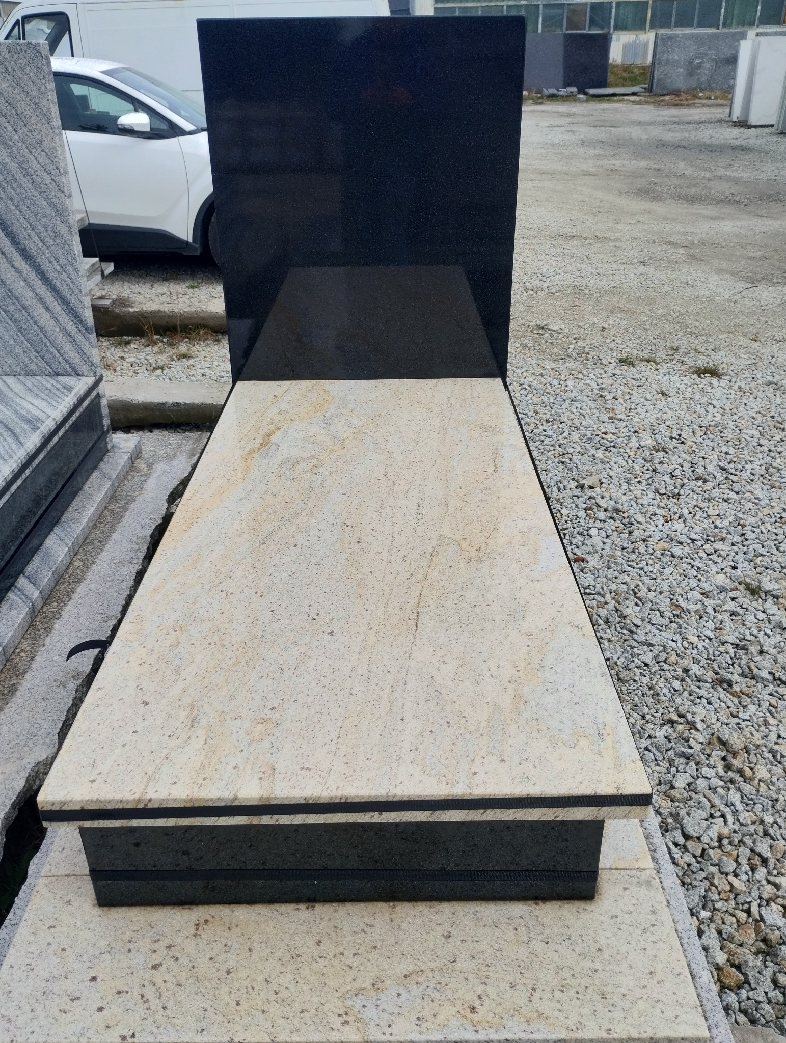 Nowoczesny nagrobek z jasnego kamienia z czarną płytą w tle, widoczny na tle placu z kamieniami.