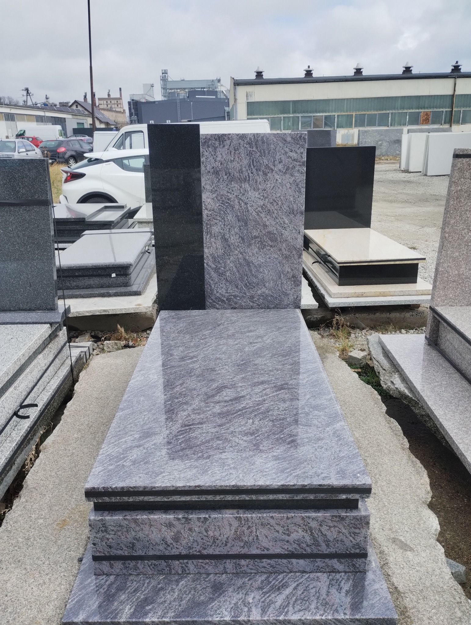 Wystawa gotowych elementów z granitu w odcieniach szarości i czerni, prawdopodobnie przeznaczonych do budowy pomników, na tle zabudowań przemysłowych i pochmurnego nieba.