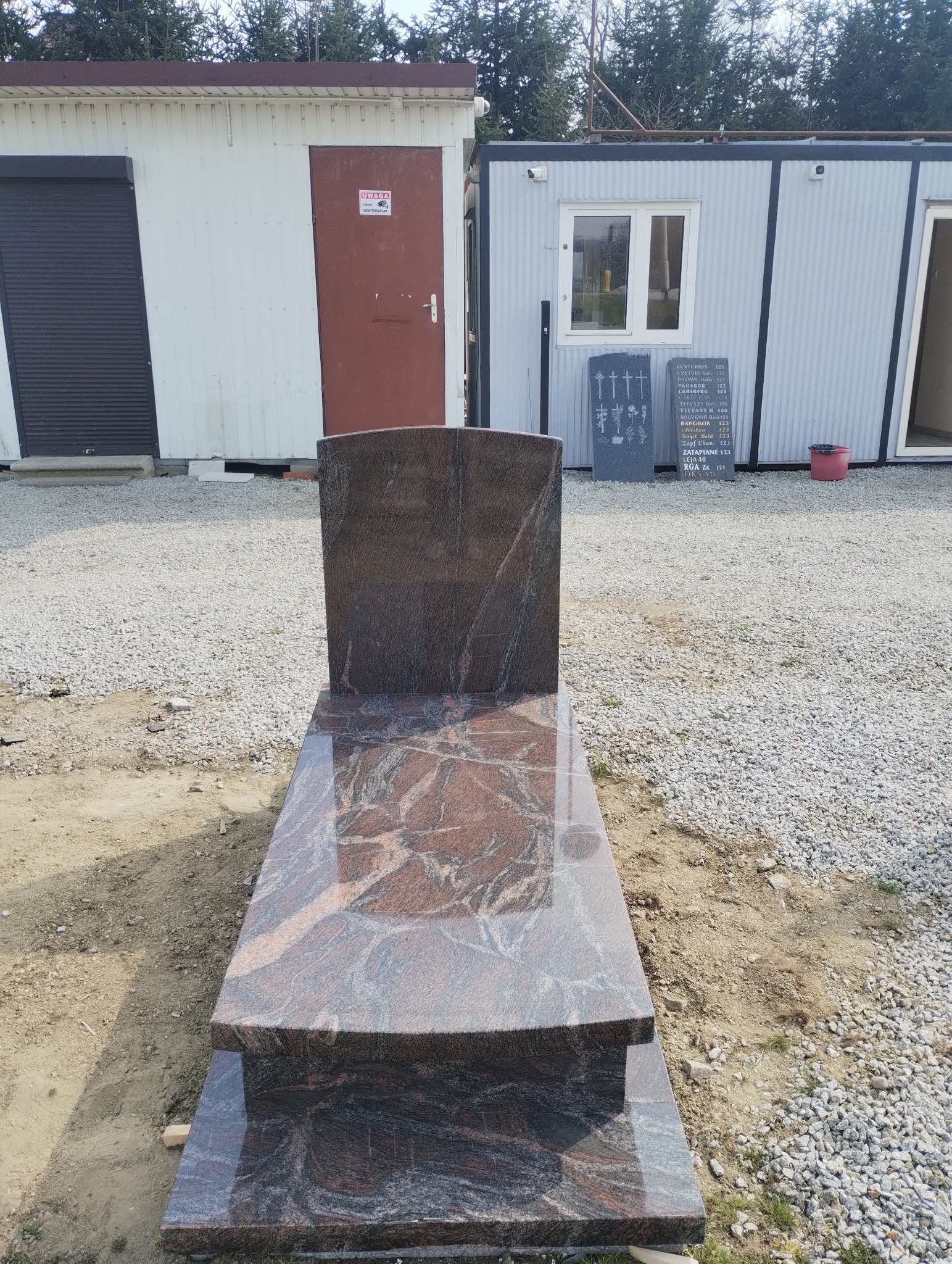 Nowy, brązowo-szary, polerowany nagrobek z granitu z widoczną strukturą kamienia, ustawiony na żwirowym placu budowy z kontenerami w tle.