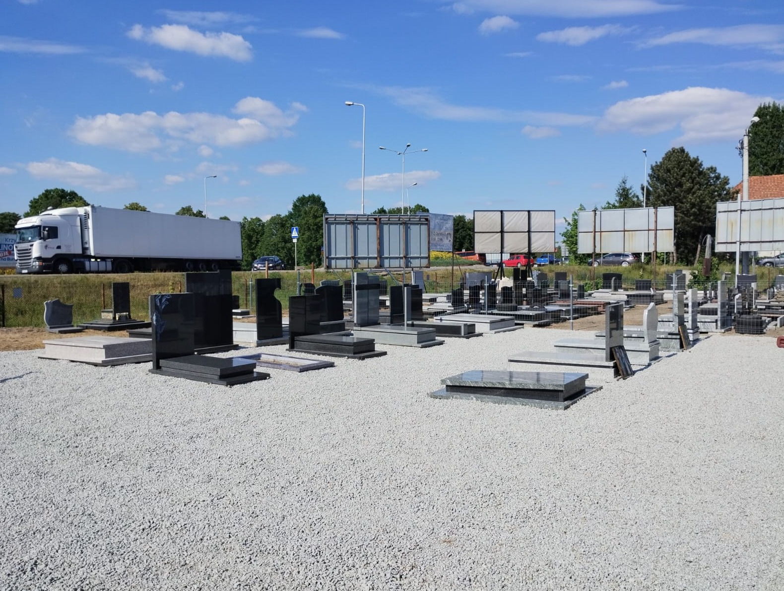 Ekspozycja różnorodnych, gotowych pomników z ciemnego i jasnego kamienia na placu z jasnym żwirem, w tle biała ciężarówka i reklamy.