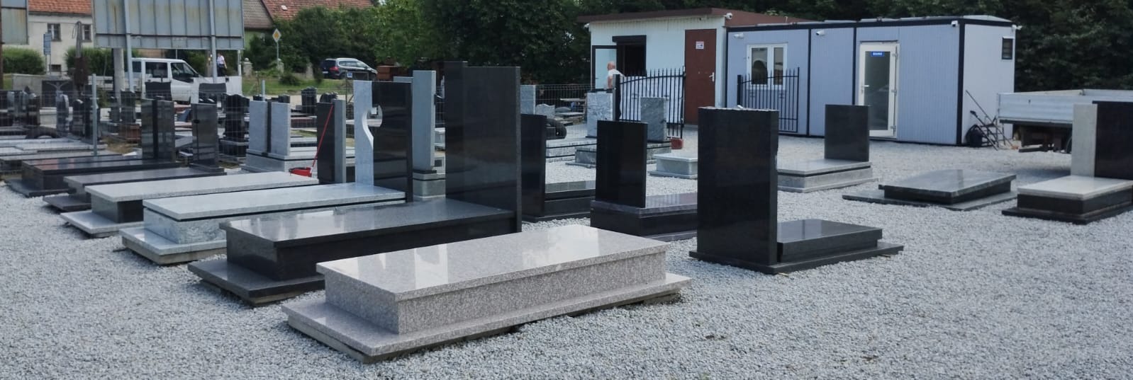 Ekspozycja różnorodnych nagrobków z granitu i marmuru, w tym modele z krzyżami i płaskie płyty, na żwirowym placu przed budynkiem biurowym i zaparkowanym samochodem dostawczym.