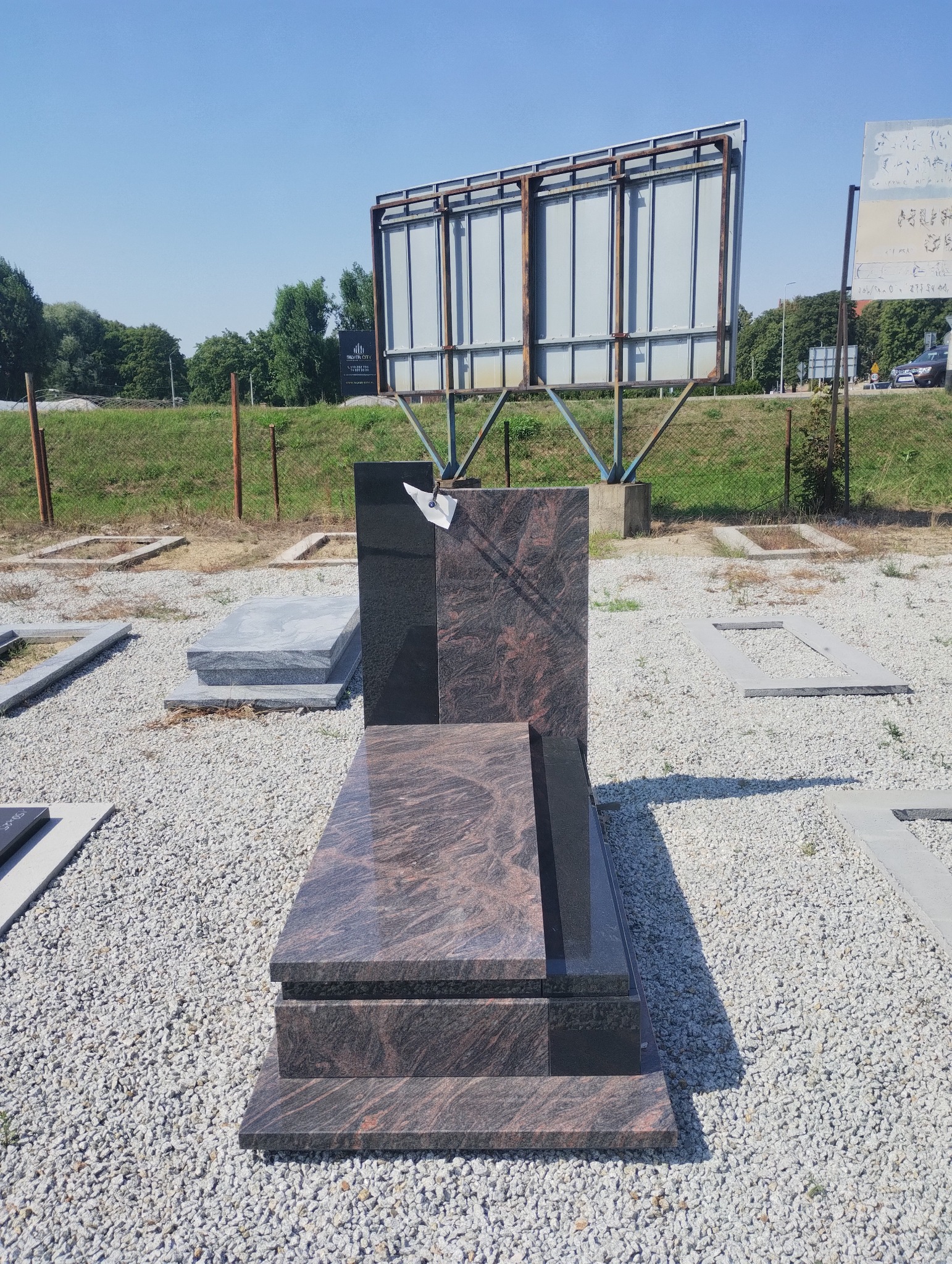 Granitowy nagrobek w odcieniach brązu i czerni, z widoczną fakturą kamienia, na tle żwirowego placu i zieleni. W tle billboard reklamowy.