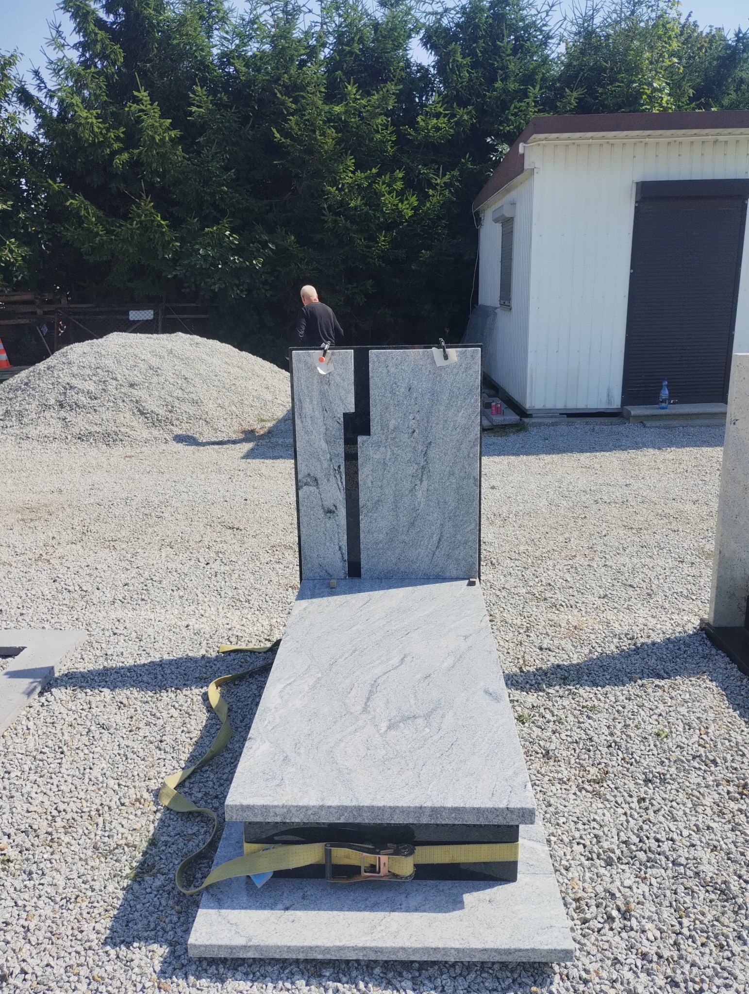 Jasnoszary nagrobek z ciemnym krzyżem, zabezpieczony pasami transportowymi na żwirowym podłożu. W tle widoczny budynek gospodarczy i kopiec żwiru.