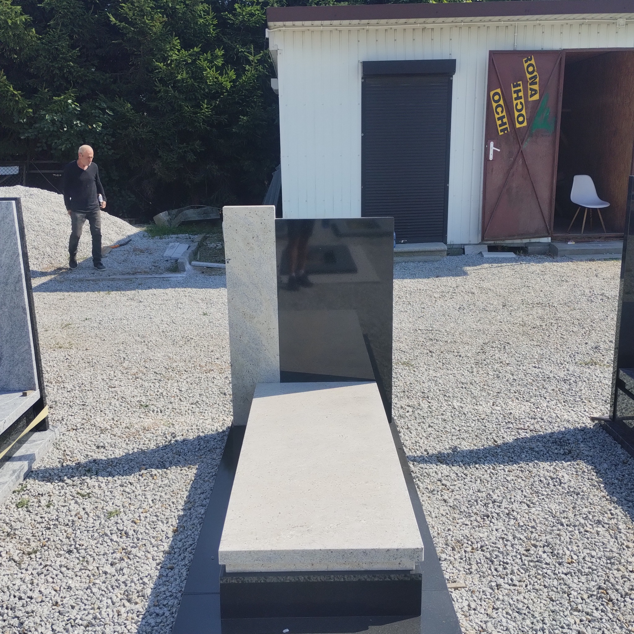 Nowoczesny nagrobek z jasnego i czarnego kamienia na żwirowym podłożu, z sylwetką mężczyzny w tle i budynkiem gospodarczym.
