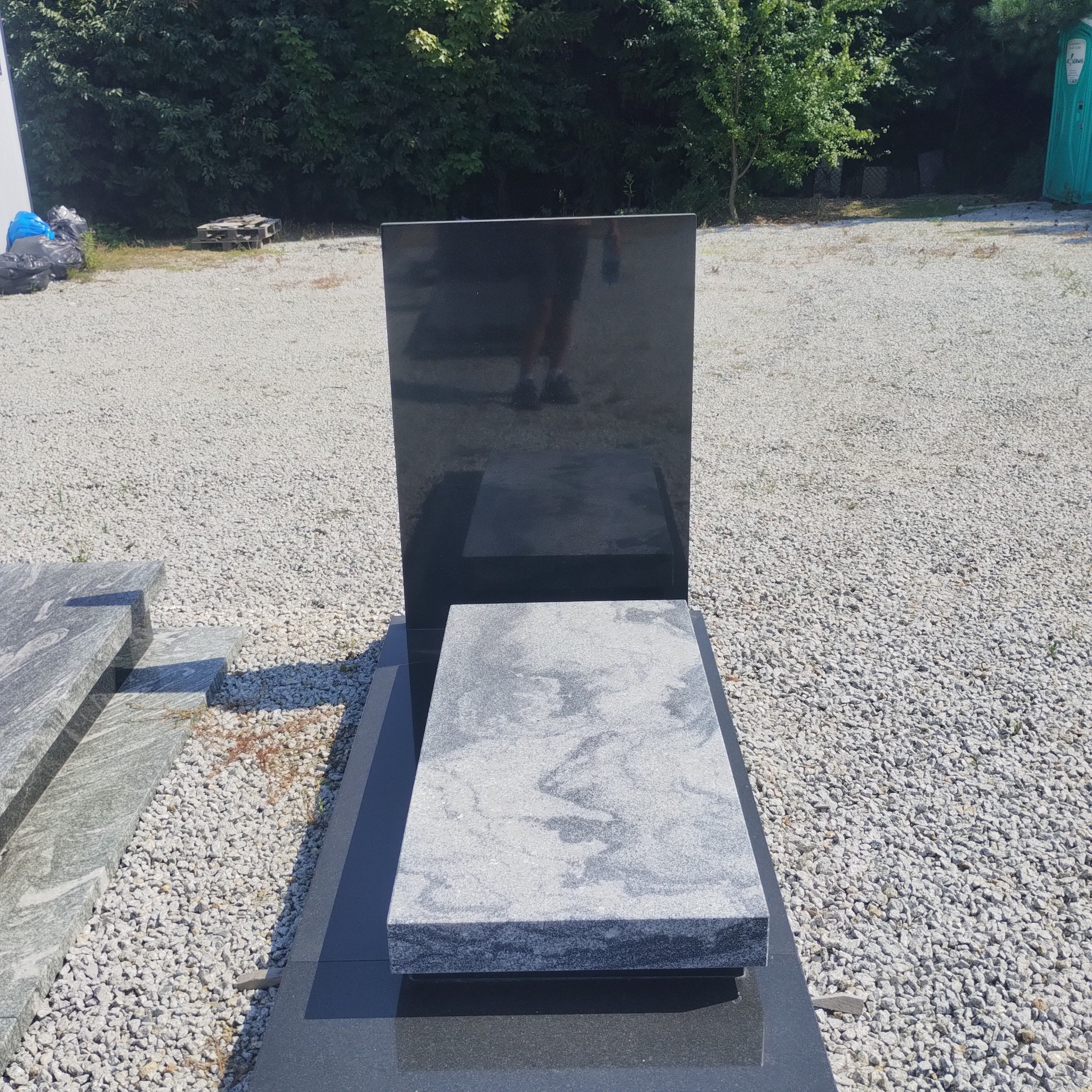 Nowoczesny nagrobek z czarnego granitu z szarą płytą wierzchnią, ustawiony na żwirowym podłożu. W tle zieleń drzew i krzewów. Widoczne odbicie osoby w polerowanej powierzchni.