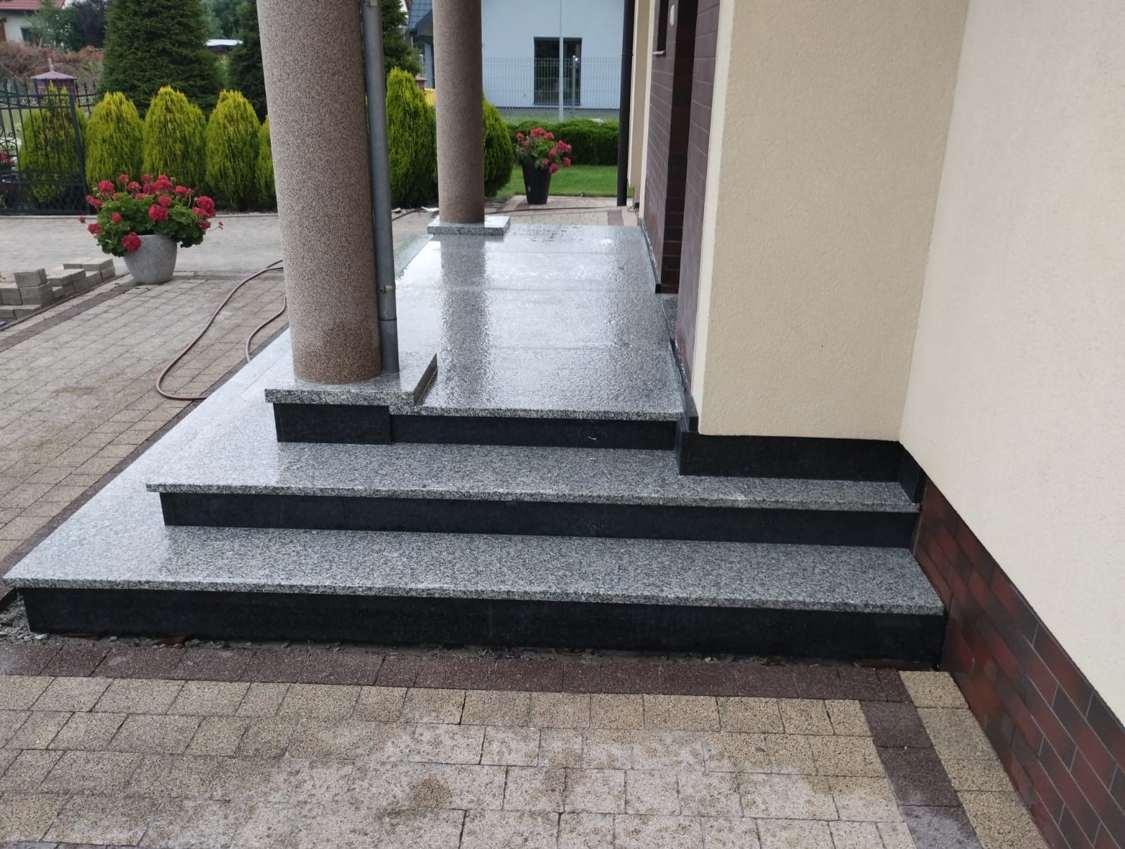 Mokre, dwukolorowe schody wejściowe z jasnoszarego i ciemnego granitu, prowadzące do pasującego podestu pod dwoma filarami. W tle zielone krzewy i dom.