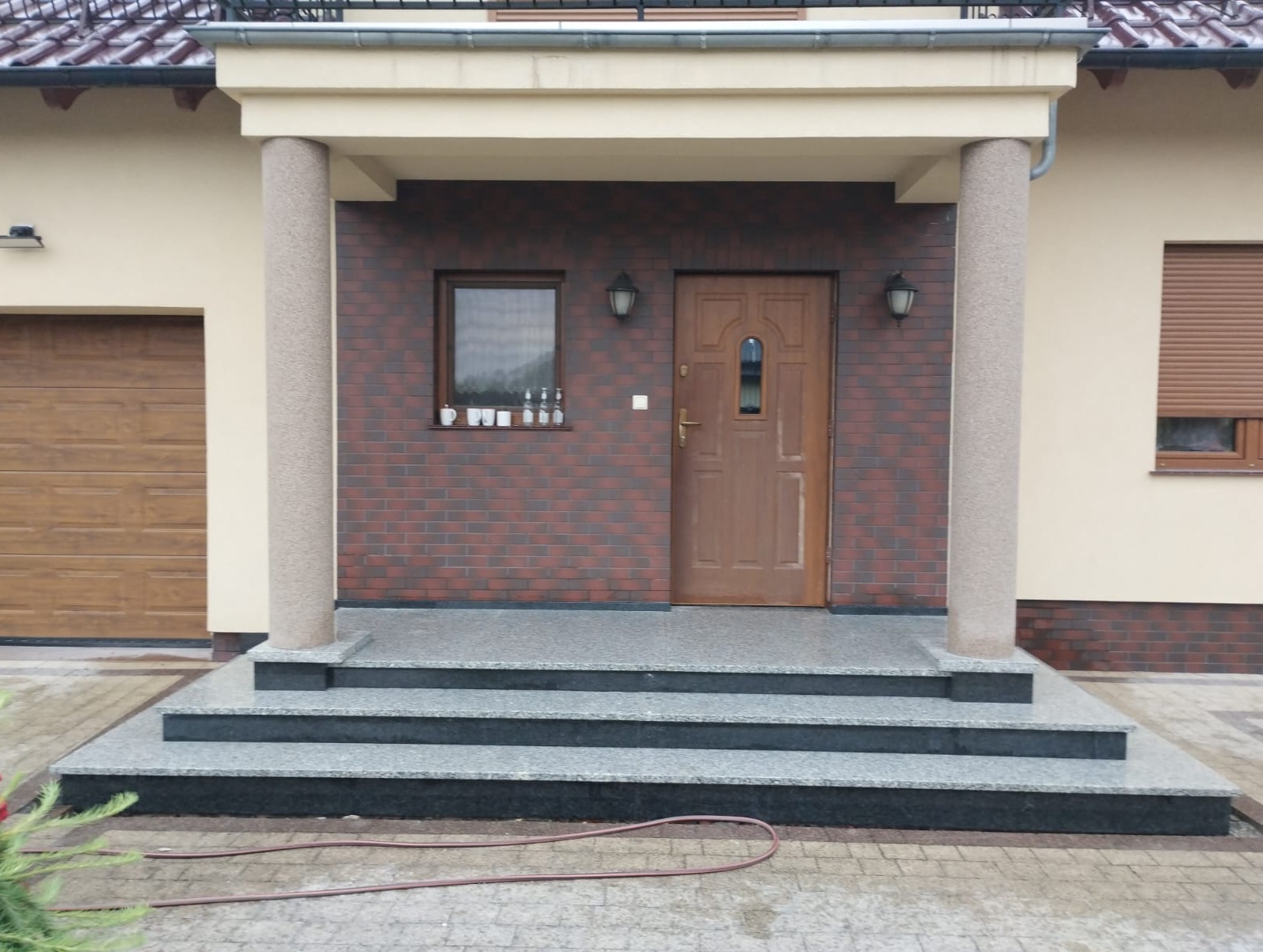 Wejście do domu z trzema stopniami z jasnoszarego kamienia i ciemnymi podstopnicami, podtrzymywane przez fakturowane beżowe kolumny. Drewniane drzwi wejściowe i ceglana elewacja.