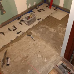 Układanie jasnych płytek podłogowych w łazience, widoczne krzyżyki dystansowe, poziomica laserowa wyznaczająca linię, paczka z klejem i zaprawa, ściany przygotowane do malowania.