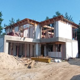 BDP Polska Daniel Budek - Budowa domu jednorodzinnego: wznoszenie ścian z bloczków, widoczne szalunki i drewniane elementy konstrukcyjne, na pierwszym planie sterta piasku i materiały budowlane, w tle las i zaparkowany bus...