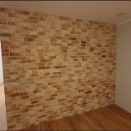 Ściana wewnętrzna wykończona dekoracyjną cegłą w odcieniach beżu i brązu, z widocznymi gniazdkami elektrycznymi, na podłodze panele drewniane.