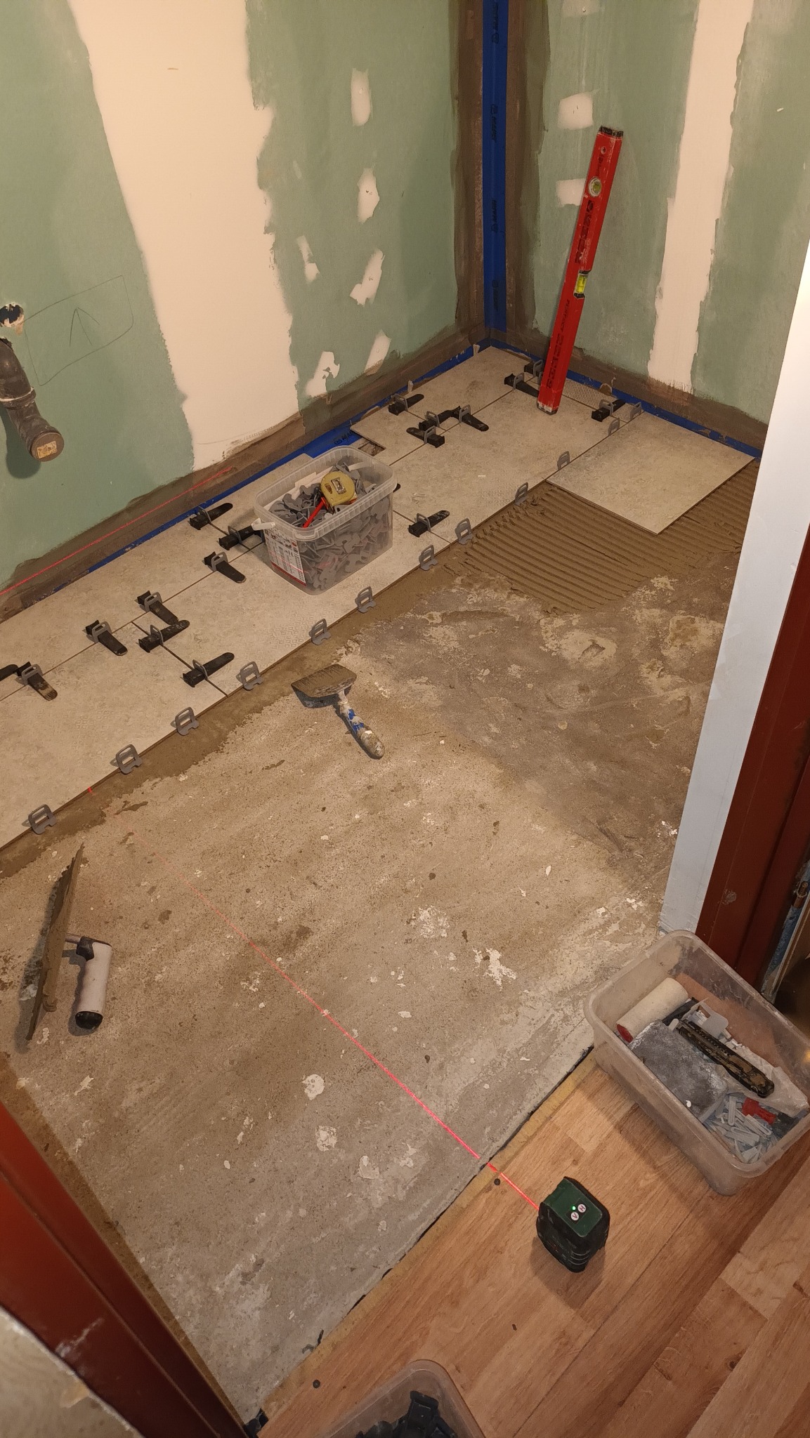Układanie jasnych płytek podłogowych w łazience, widoczne krzyżyki dystansowe, poziomica laserowa wyznaczająca linię, paczka z klejem i zaprawa, ściany przygotowane do malowania.
