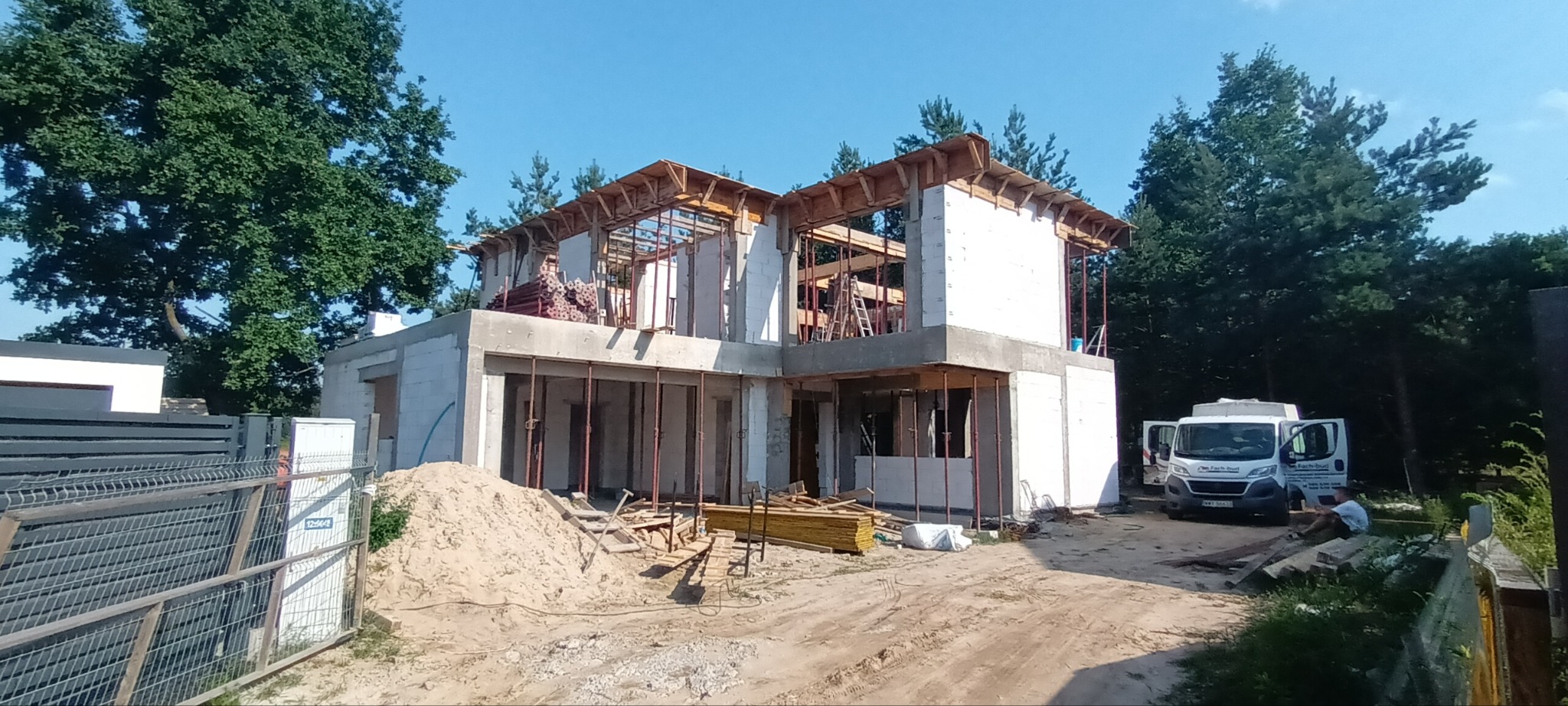 Budowa domu jednorodzinnego: wznoszenie ścian z bloczków, widoczne szalunki i drewniane elementy konstrukcyjne, na pierwszym planie sterta piasku i materiały budowlane, w tle las i zaparkowany bus...