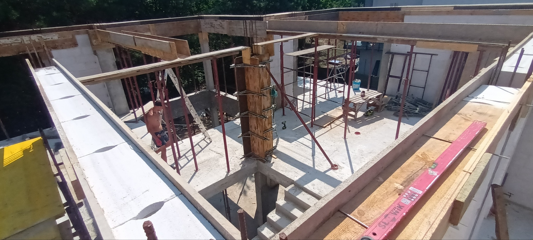 Wysoki kąt widzenia na plac budowy z widocznymi szalunkami drewnianymi, rusztowaniami i pracownikiem w tle, ukazujący etap wznoszenia konstrukcji budynku.