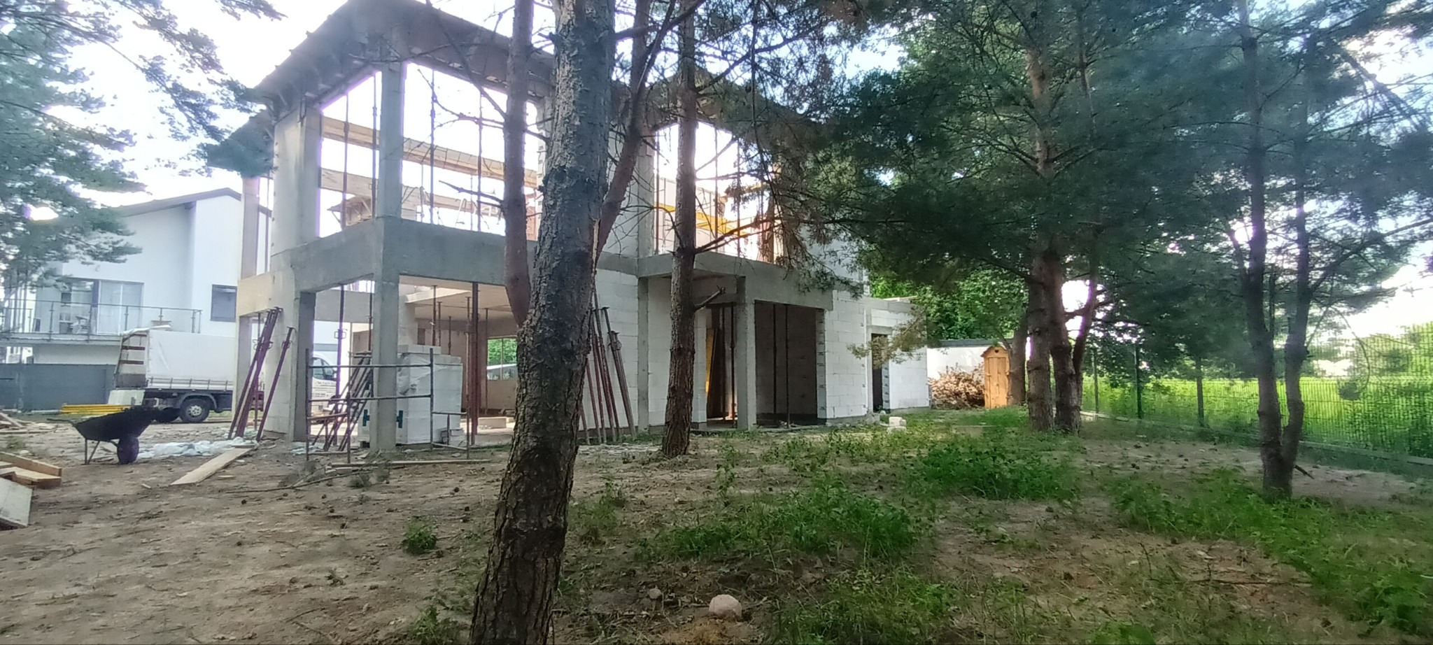 Budowa domu jednorodzinnego w stanie surowym otwartym, widoczne białe bloczki i betonowe słupy, na pierwszym planie drzewa iglaste, w tle drugi dom i samochód dostawczy.