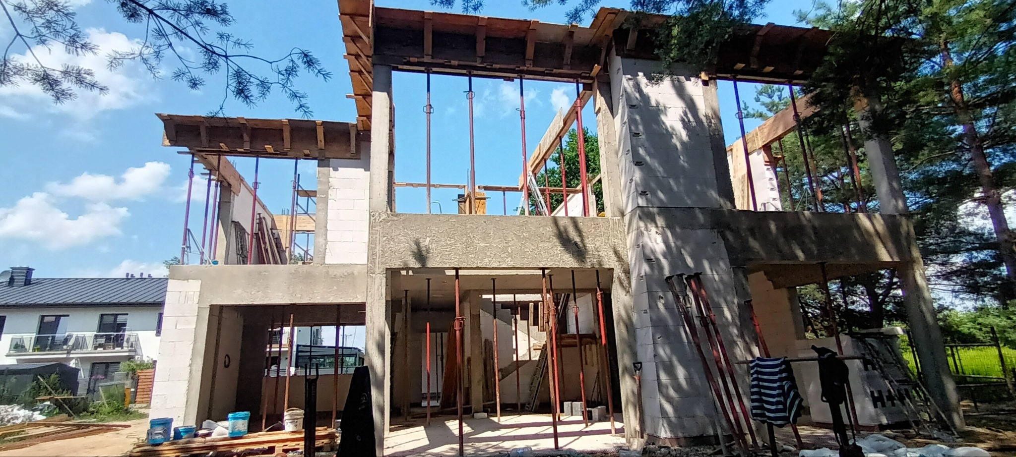 Surowy stan dwukondygnacyjnego domu w trakcie budowy z widocznym szalunkiem, podporami i częściowo wymurowanymi ścianami z betonu komórkowego na tle błękitnego nieba i drzew.