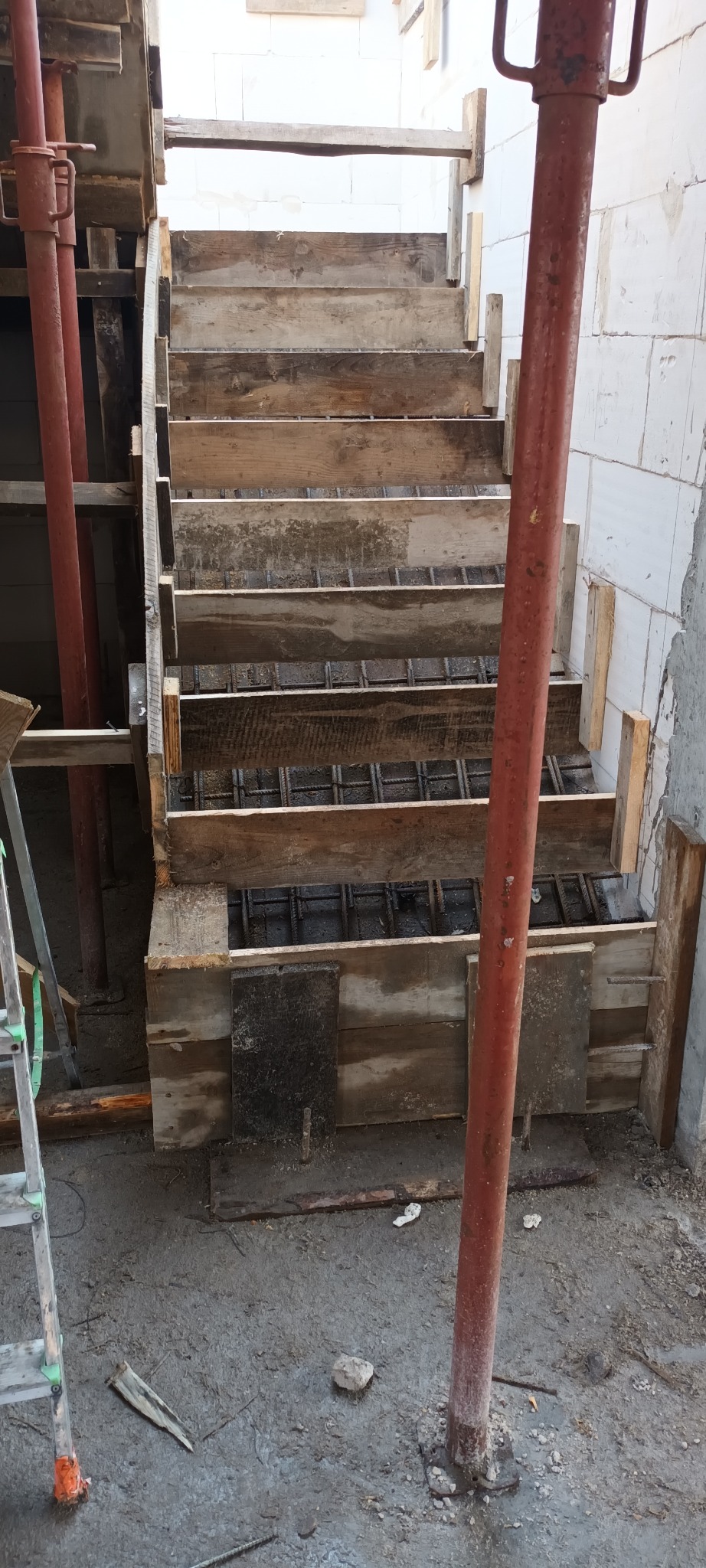 Szare, betonowe schody w trakcie budowy, z widocznym zbrojeniem i drewnianym szalunkiem, podparte metalowymi stemplami.