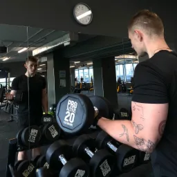 Mężczyzna z tatuażami na ramieniu ćwiczy biceps z hantlą 20 kg, widoczny w lustrze w tle siłowni z innymi hantlami i sprzętem do ćwiczeń.
