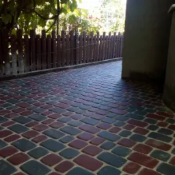 Ułożona kostka brukowa w odcieniach czerwieni i szarości na podw&oacute;rku, widok z bliska na wz&oacute;r ułożenia, drewniany płot w tle, częściowo zacienione otoczenie.