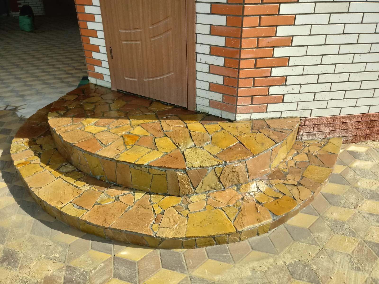 Półokrągłe schody wejściowe wyłożone nieregularnymi, żółto-brązowymi płytkami kamiennymi, prowadzące do drewnianych drzwi z ceglaną obudową.