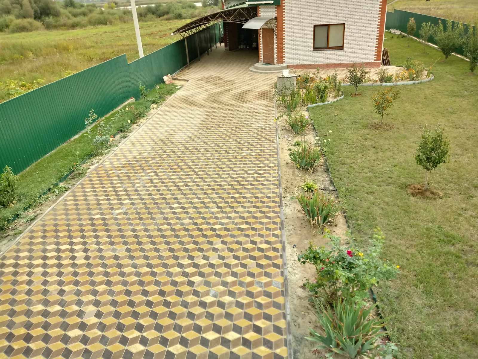 Ułożona kostka brukowa w geometryczny wzór 3D w kolorach żółtym, brązowym i czarnym prowadząca do domu z białej cegły, obok zielony trawnik i ogrodzenie.