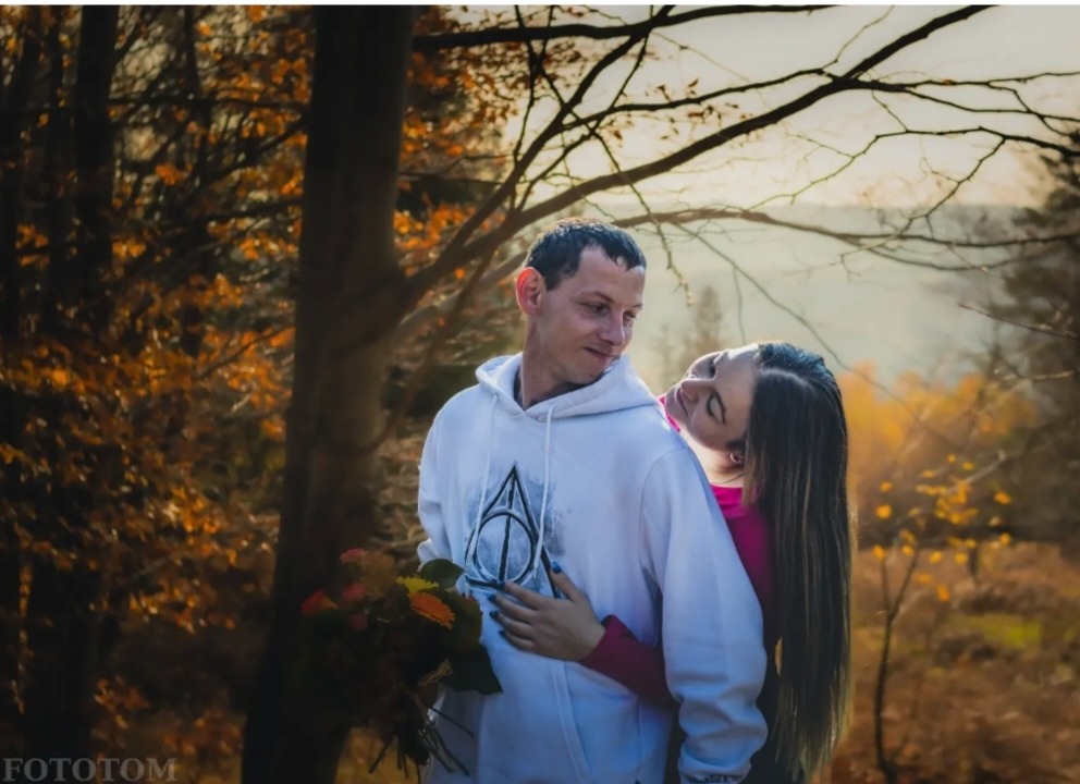 Para w jesiennym lesie: mężczyzna w białej bluzie z kapturem, kobieta opiera głowę o jego ramię, trzymając bukiet kwiatów.