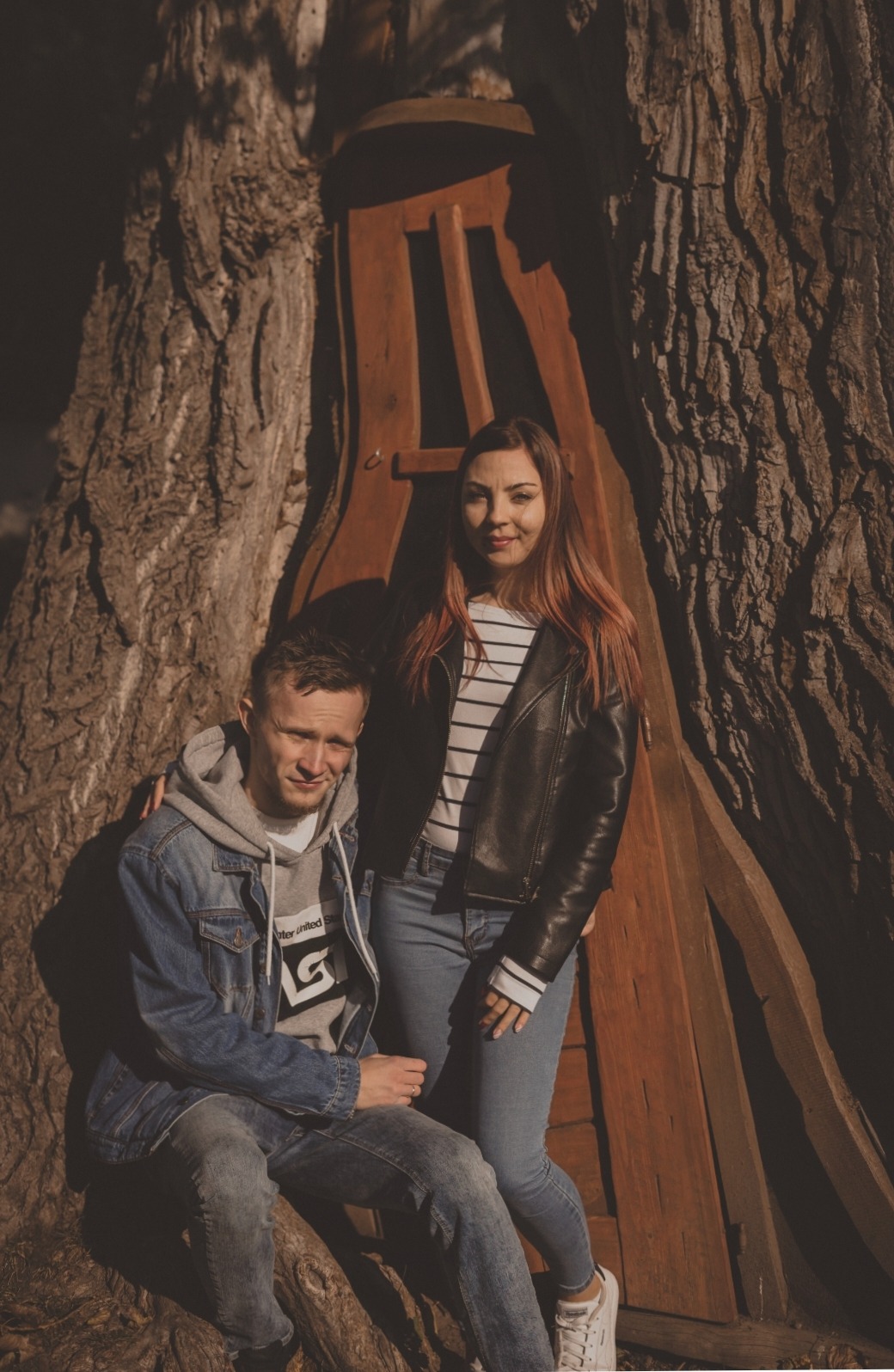 Para pozuje do zdjęcia przy dużym drzewie z nietypową drewnianą konstrukcją przypominającą drzwi, on siedzi w dżinsowej kurtce i bluzie z kapturem, ona stoi w skórzanej kurtce i dżinsach.