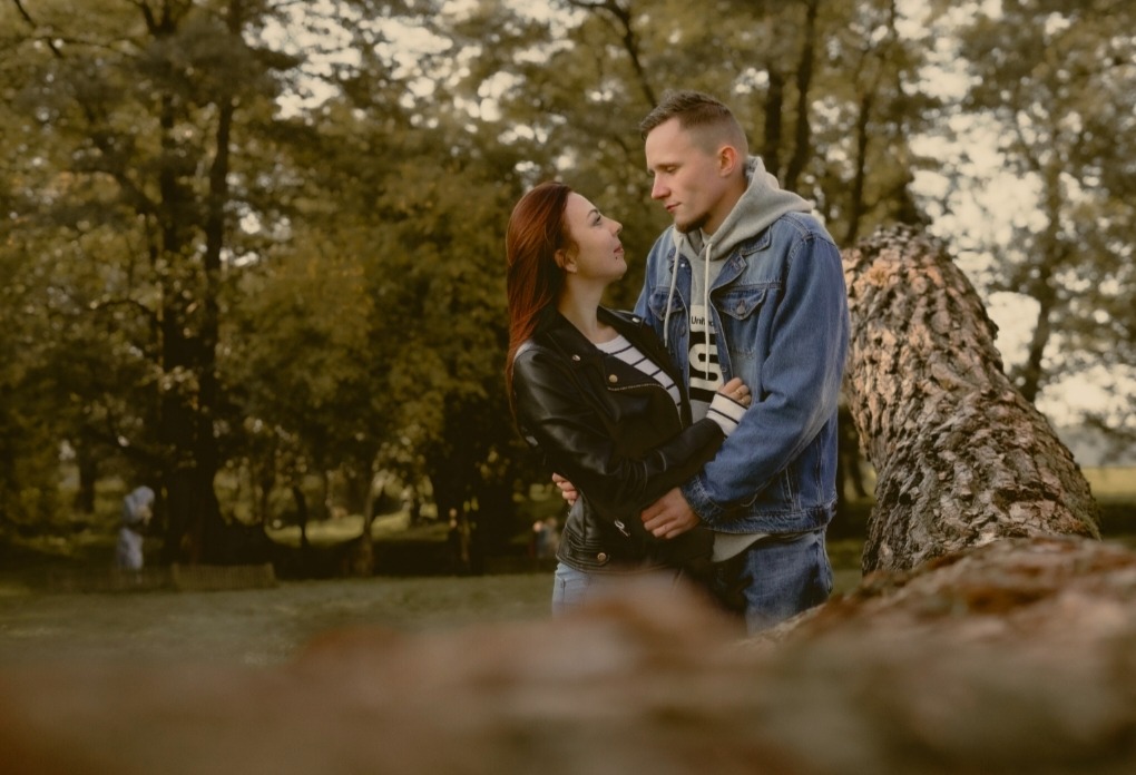 Para obejmująca się w parku, kobieta w skórzanej kurtce patrzy na mężczyznę w jeansowej kurtce, w tle drzewa i powalone pnie.