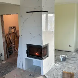 Wnętrze w trakcie wykańczania z kominkiem obłożonym białymi płytami imitującymi marmur, widoczny płomień w palenisku, podłoga zabezpieczona folią, drabina w tle.