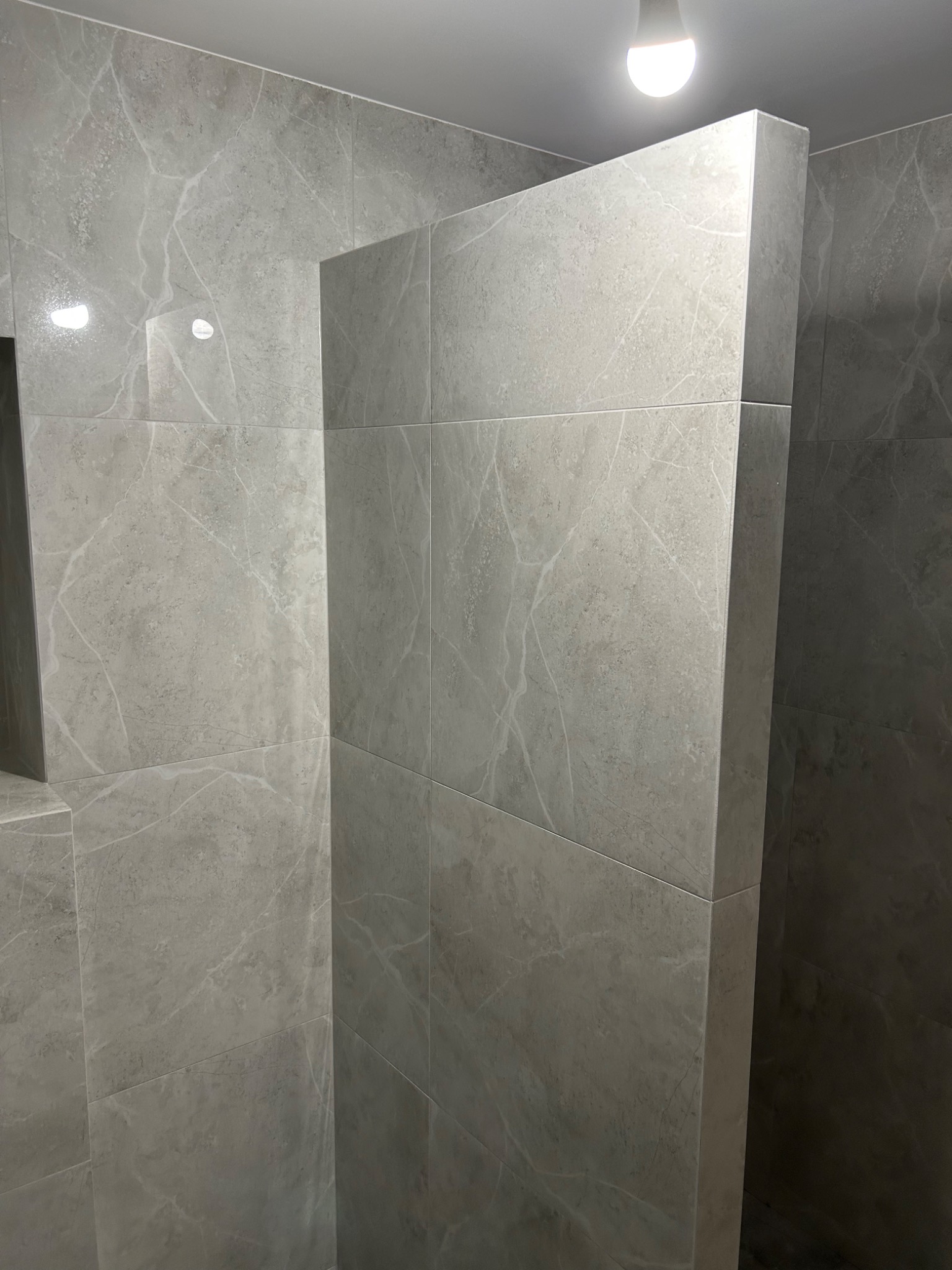 Wykończona łazienka z szarymi płytkami imitującymi marmur na ścianach, oświetlona pojedynczą żarówką sufitową.