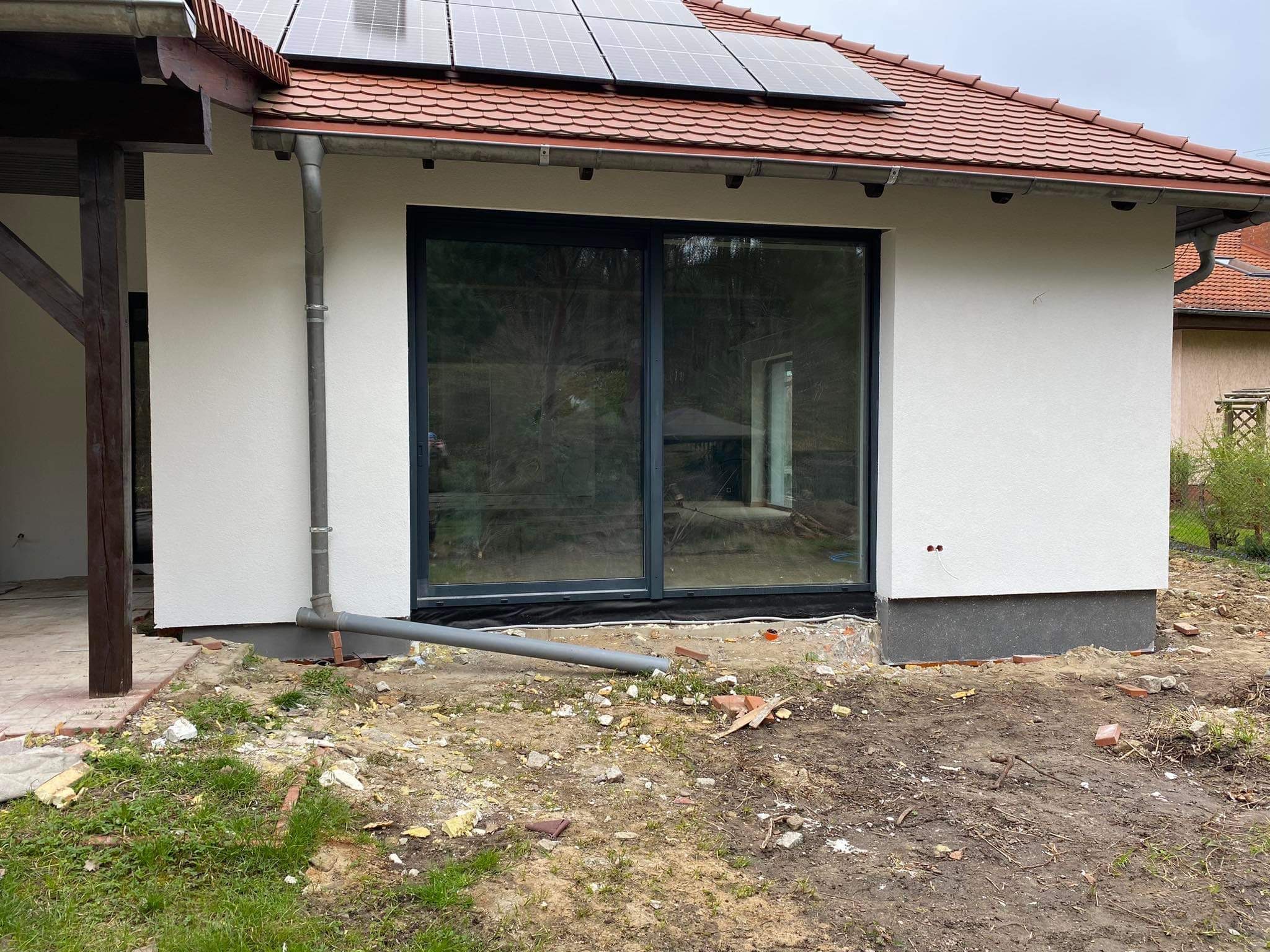 Nowa, biała elewacja domu jednorodzinnego z szarymi oknami przesuwnymi i panelami słonecznymi na dachu, widoczny stan surowy wokół budynku.