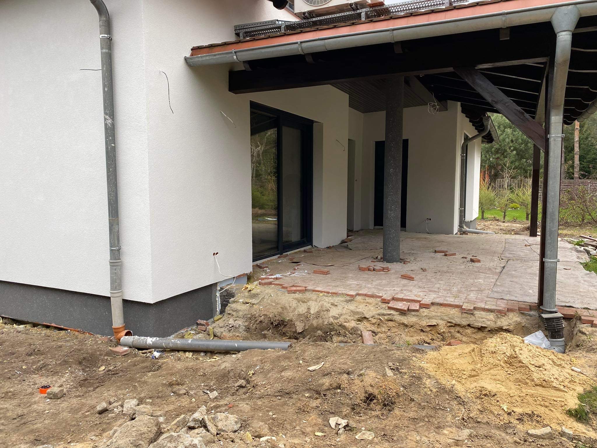 Przygotowania do prac ziemnych przy elewacji domu z wykopanymi fundamentami, częściowo ułożonymi cegłami, rurami odprowadzającymi wodę i świeżo nałożoną, jasną elewacją.