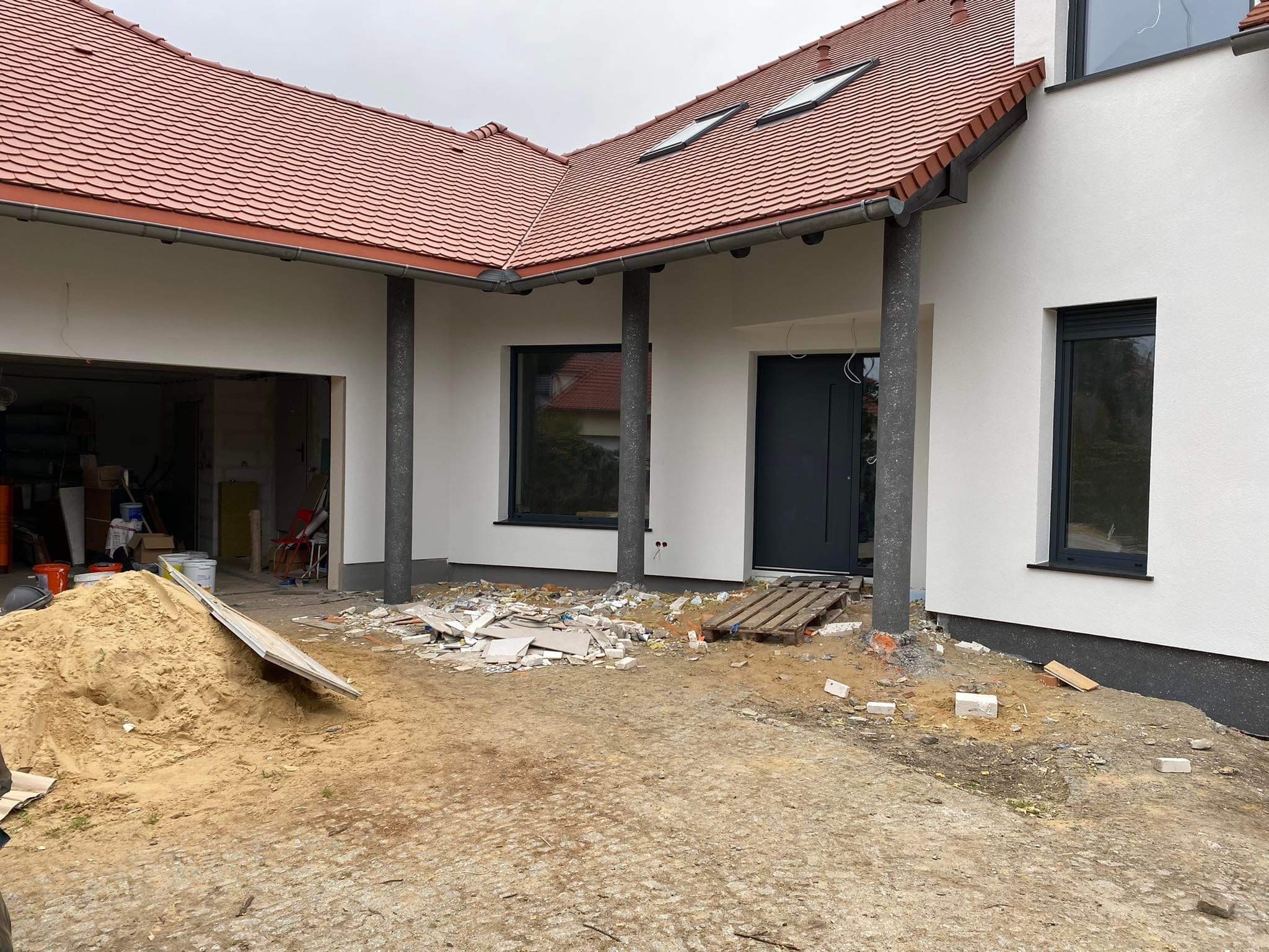 Dom jednorodzinny w trakcie budowy, widoczna elewacja z oknami i drzwiami, sterta piasku i gruzu przed wejściem, czerwona dachówka.